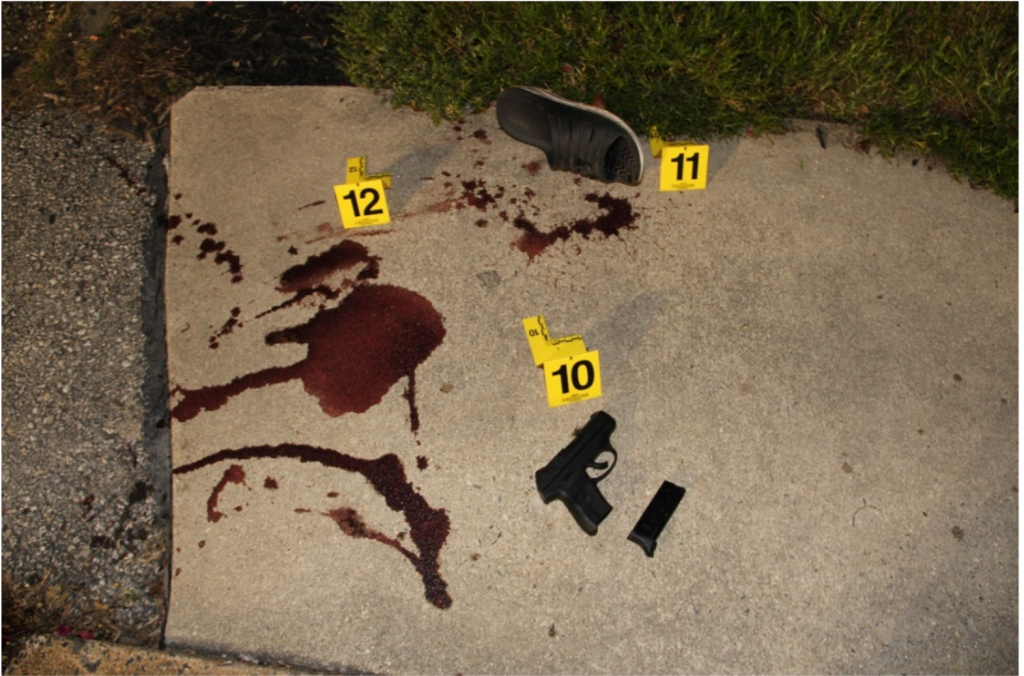 A handgun, blood stains, and a shoe on a sidewalk with evidence exhibit markers.